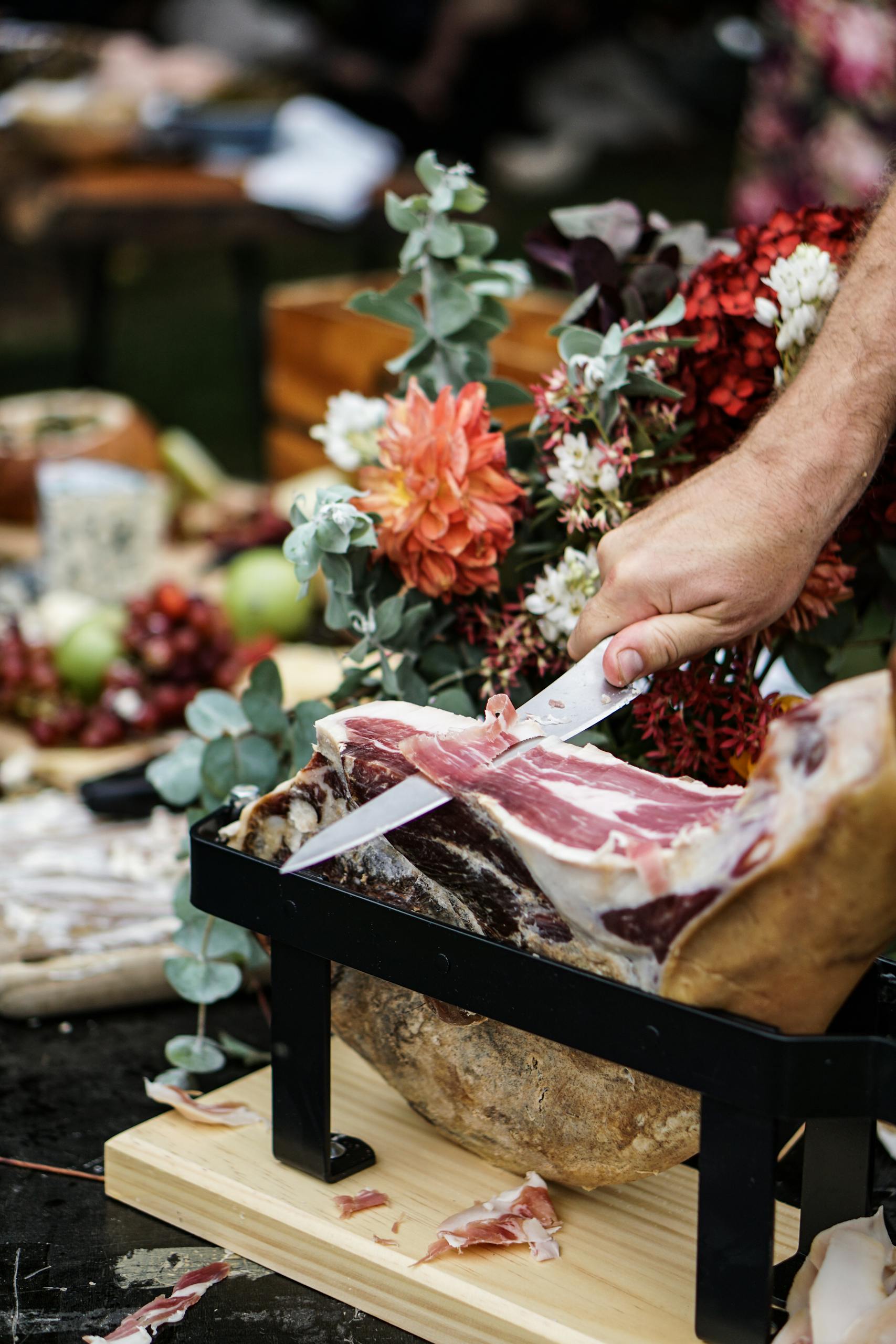 A hand skillfully carves ham surrounded by vibrant flowers and fruits.