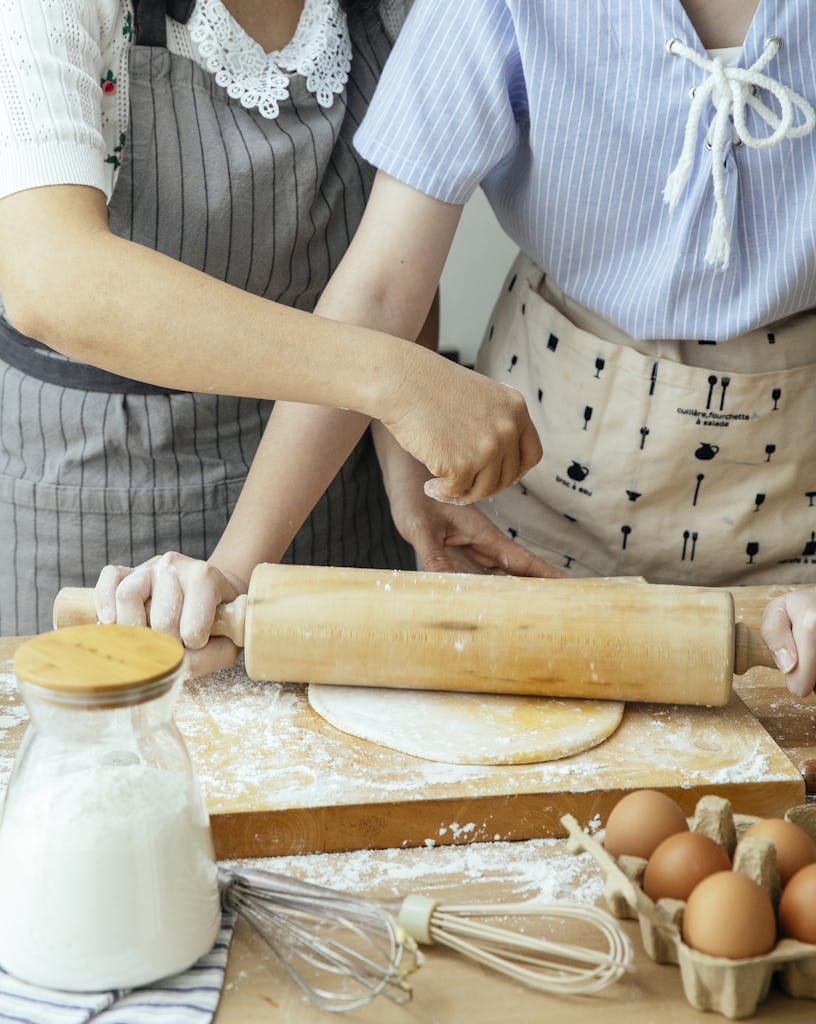 Two women in aprons baking in a home kitchen, rolling dough together.